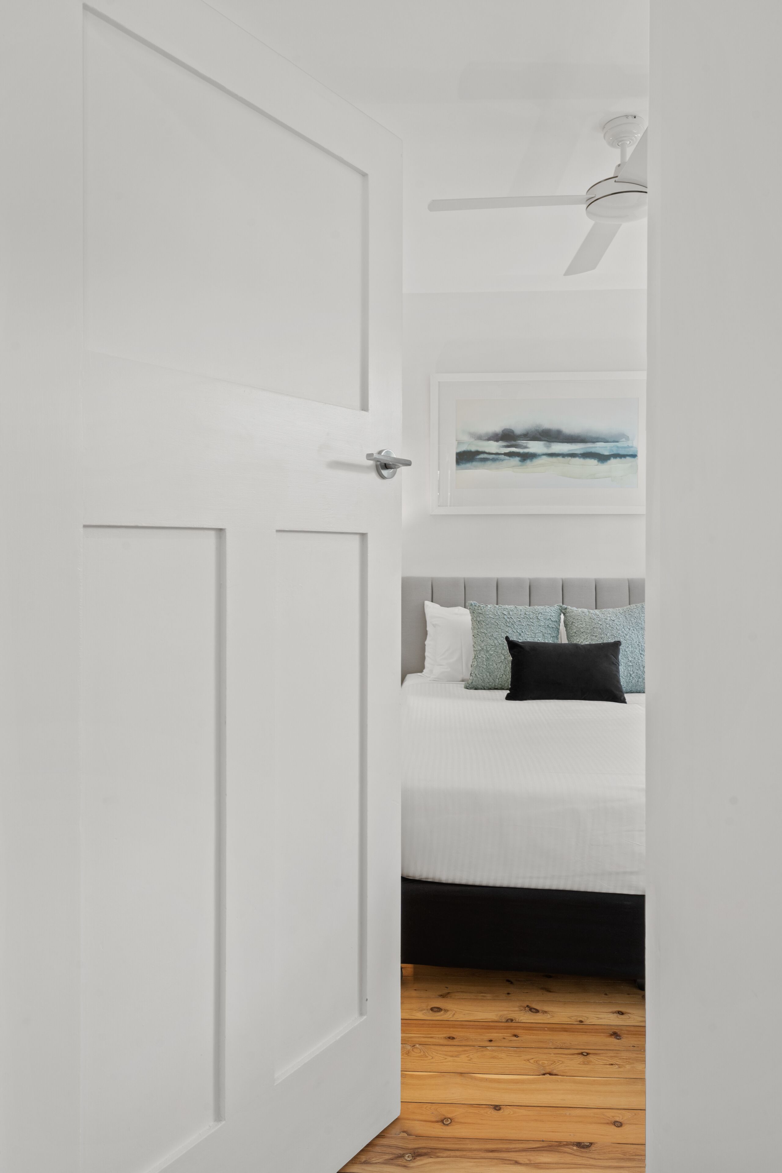 Een slaapkamer met een witte deur, een plafondventilator, een bed met een grijs hoofdeinde en een ingelijst schilderij aan de muur.