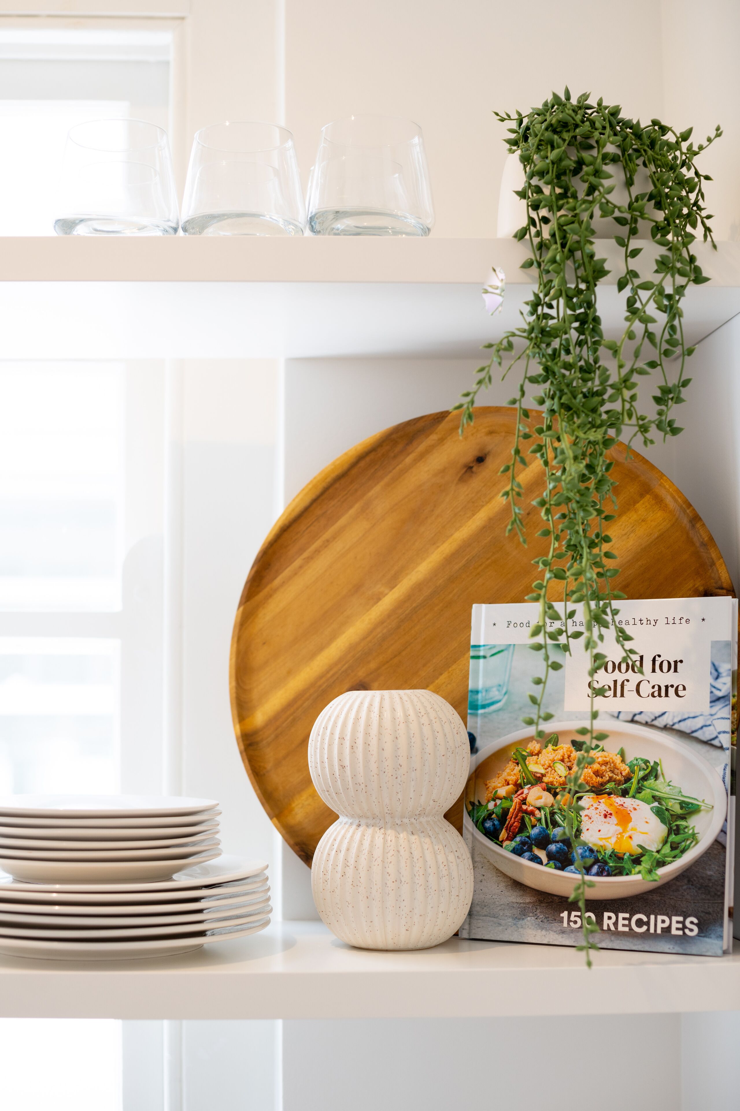 A kitchen counter with a stack of white plates, a wooden bowl, a white vase, a bowl of salad, and a cookbook titled "Food for Self-Care" with 150 recipes.