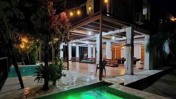 Et indendørs poolområde med en pool oplyst i grønt, omgivet af loungesæder og en overdækket terrasse.