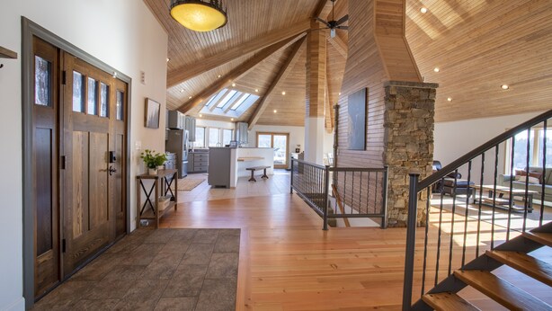 Un interior espacioso con piso de madera, una escalera con un pilar de piedra y vista a la cocina a través de un techo abierto.