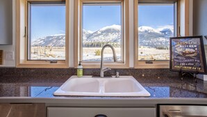 Una cocina con un lavamanos doble, tres ventanas con vistas a la montaña y un letrero que anuncia una tarta de caballa.