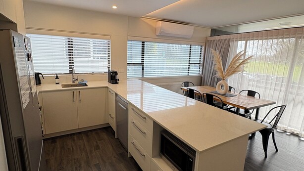A modern kitchen with white cabinetry, a central island, and a dining area with a table and chairs. - Location Location! T Two on Whakaue  (Rotorua)