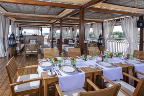 Un área de comedor al aire libre con mesas y sillas de madera, cubierta por una pérgola de madera, con vista a un cuerpo de agua.