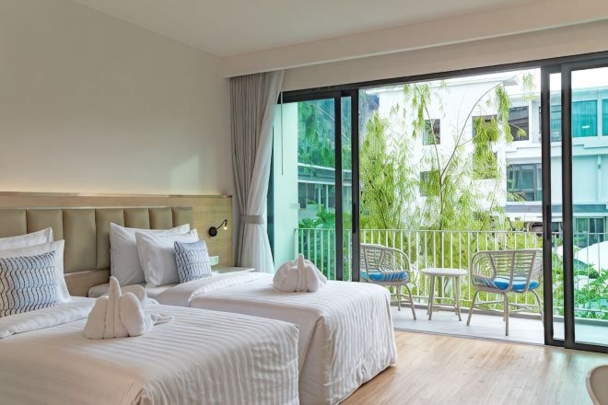 A hotel room with two beds, a balcony with a view of greenery and buildings, and a dining area with chairs.
