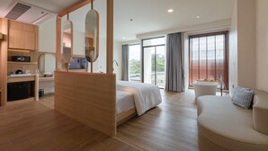 A modern hotel room with a wooden bed, a sitting area with a sofa, a small kitchen with a refrigerator, and a large window with blinds. - Infinity Aonang Krabi Hotel (Krabi)