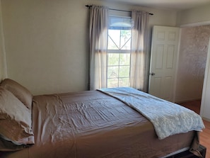 A bedroom with a single bed, a window with curtains, a door, and a plain wall.