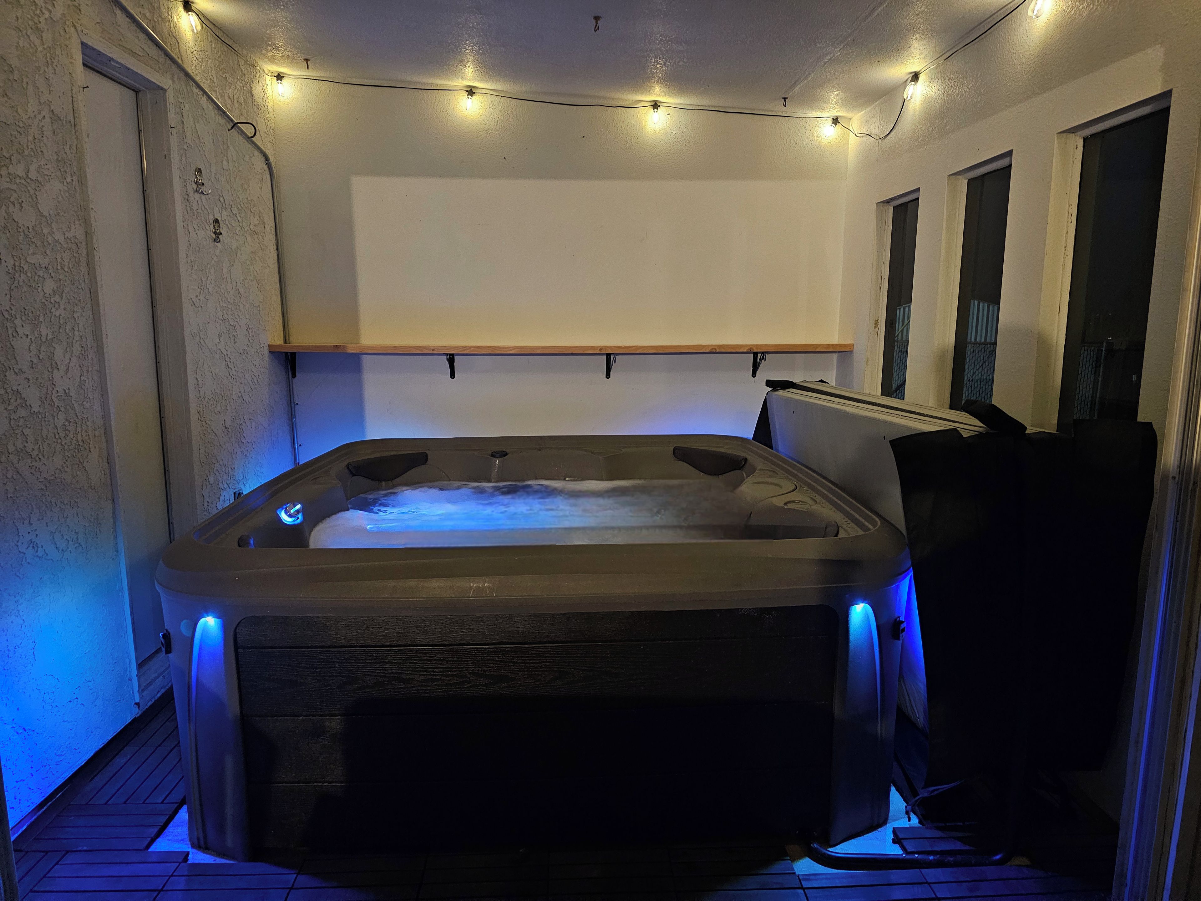 Una vasca idromassaggio con una panchina di legno, circondata da un pavimento piastrellato e una ghirlanda di luci sopra.