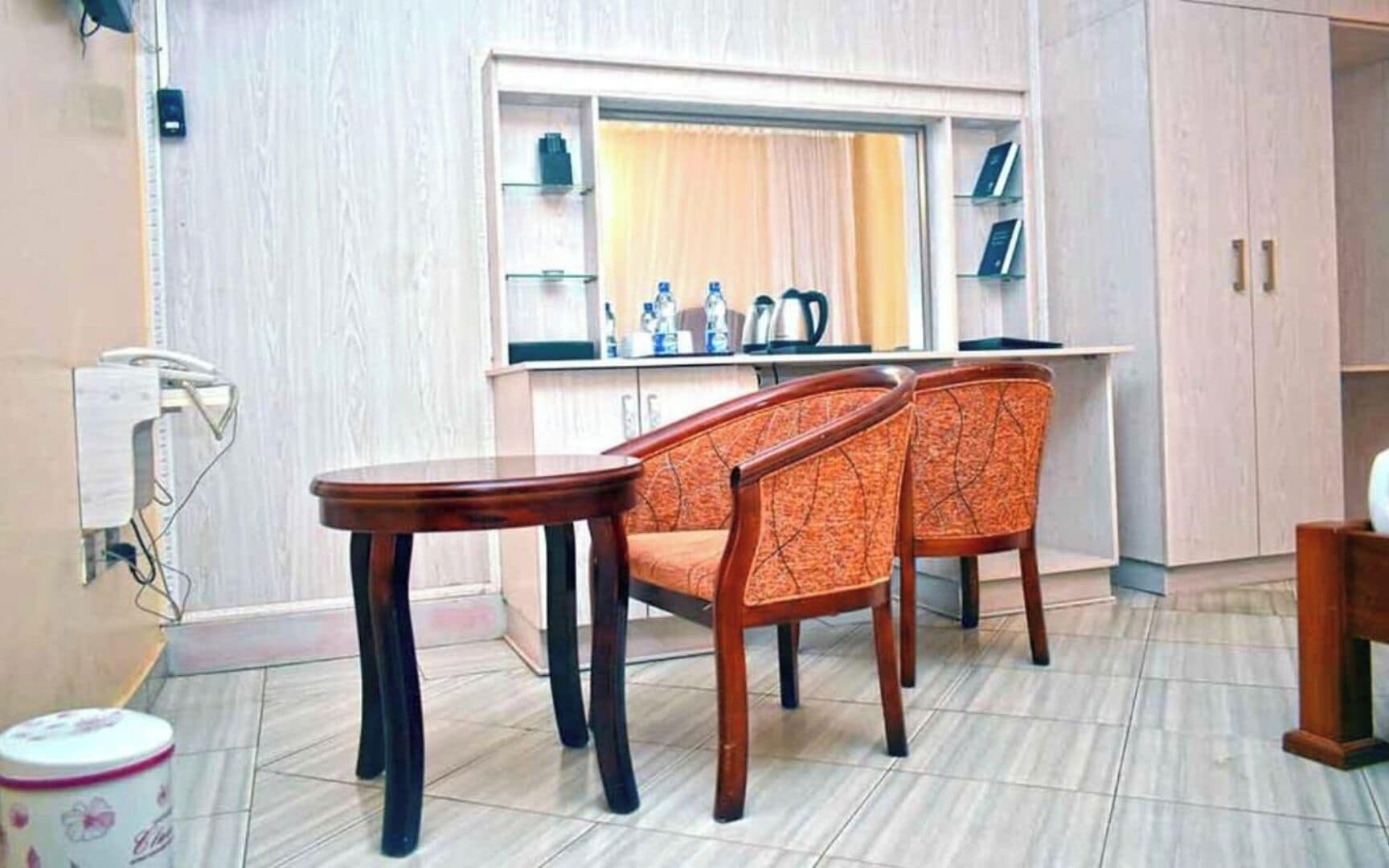 A room with a dining table and two chairs, a small cabinet with kitchen items, and a window with curtains.