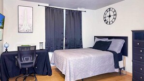 Ein Schlafzimmer mit einem Bett, einem Schreibtisch, einem Stuhl, einer Kommode, einer Uhr und einem Fenster mit Vorhängen.