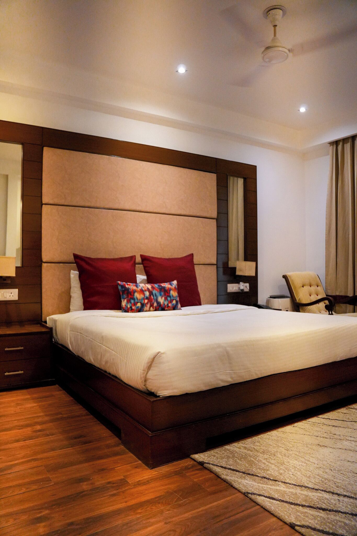 Ein Hotelzimmer mit einem großen Bett, einem Holzkopfteil, Nachttischen, einem Sessel und einem Deckenventilator.