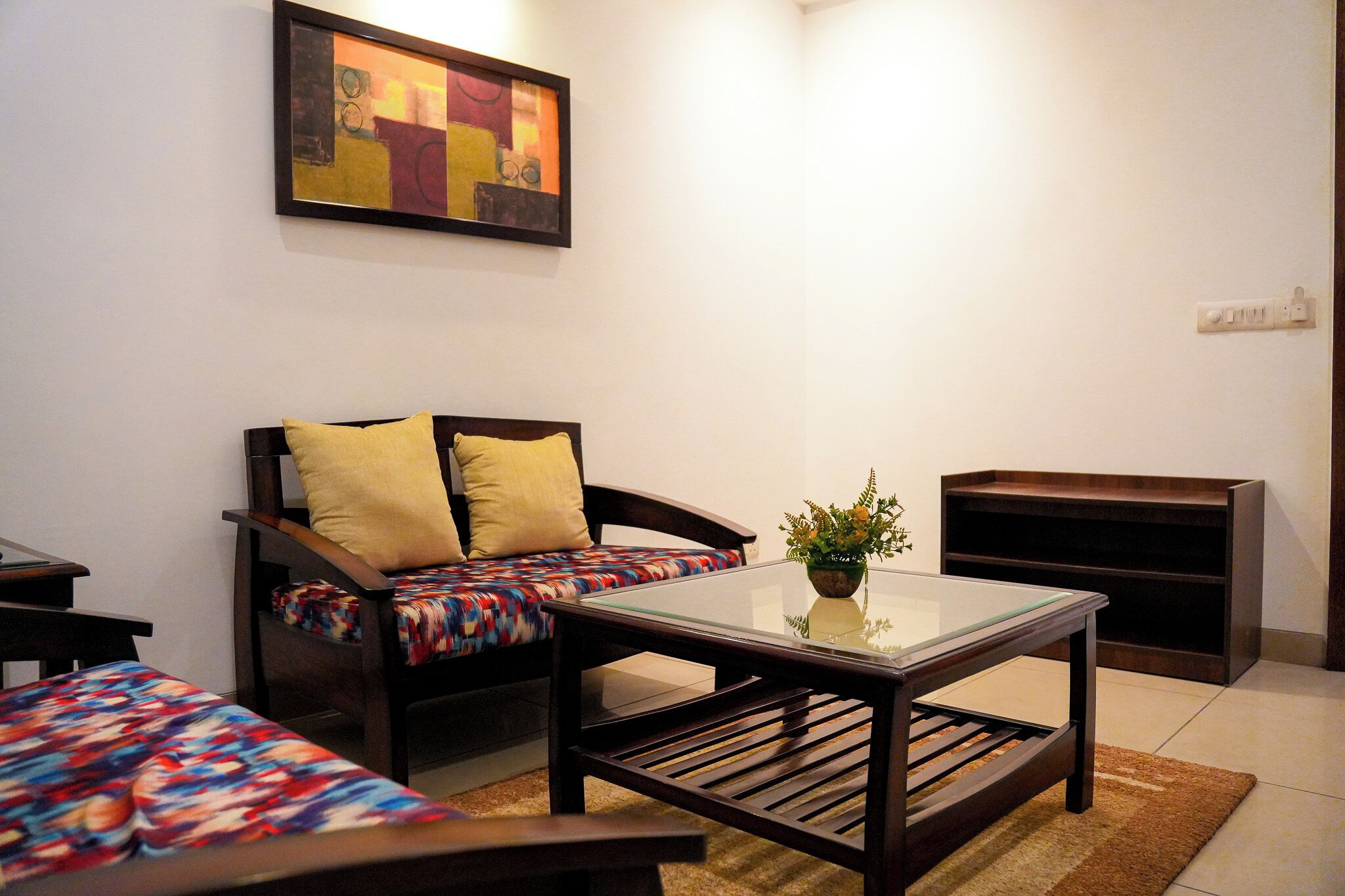 A small, furnished room with a sofa, a glass-top table, a bookshelf, and a painting on the wall.