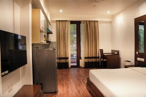 A hotel room with a bed, a desk, a refrigerator, a television, and a view of the outdoors through a door. - Yuhi Suites (Gurugram)