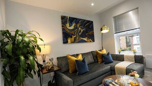 Un salon moderne avec un canapé gris, un grand tableau abstrait, une plante en pot et une table basse sur laquelle sont posés des livres et une corbeille de fruits.