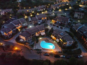 An aerial view of a residential area with houses, a swimming pool, and a well-lit street at dusk.