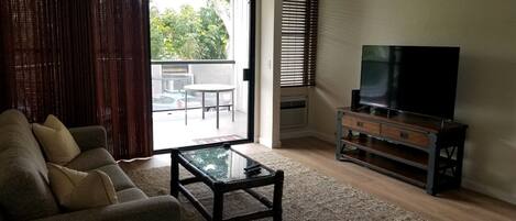 Un salón con sofá, una mesa de centro, un televisor sobre un mueble de madera y vistas al exterior a través de un ventanal corredero.