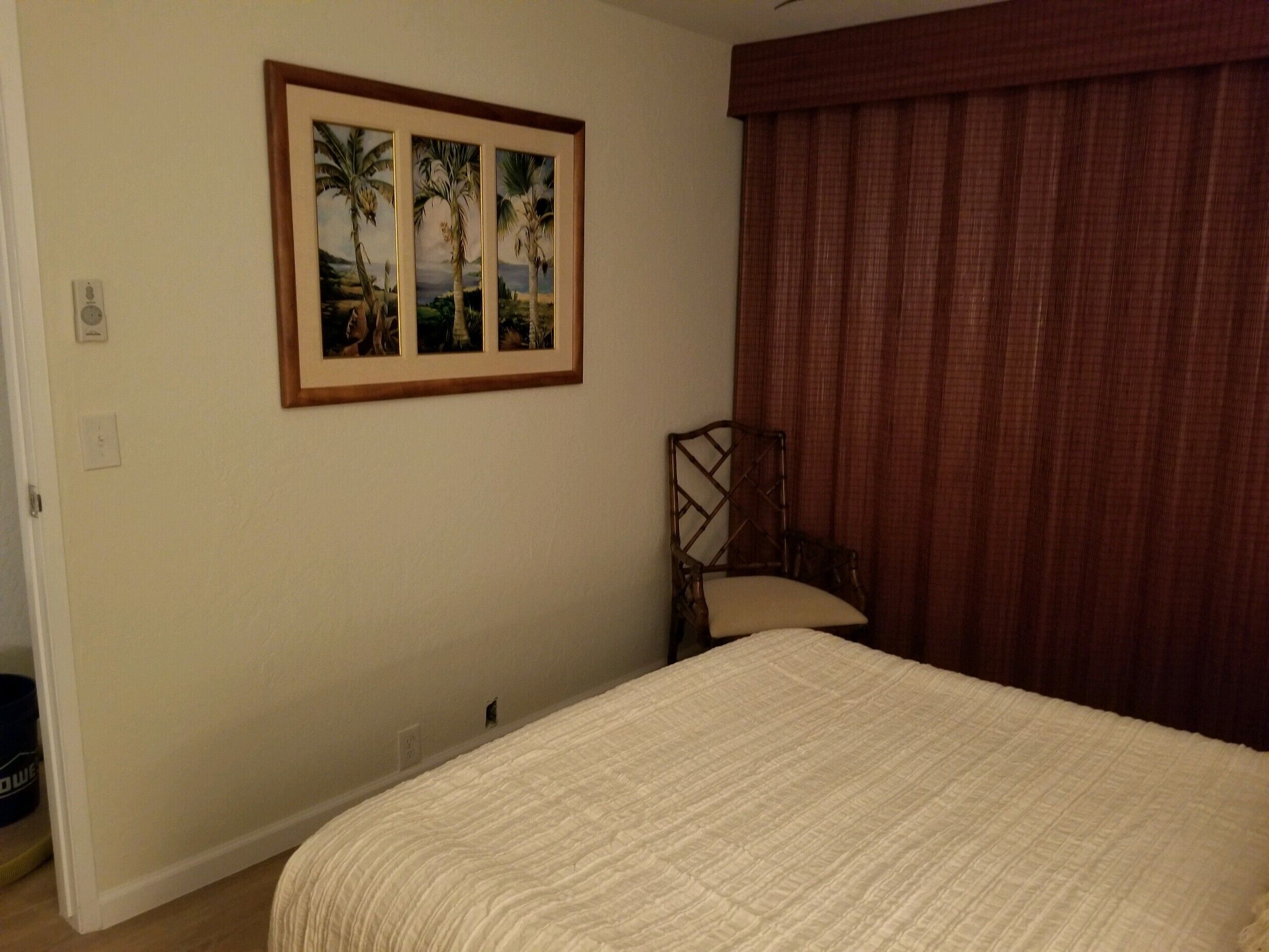 Un dormitorio con cama, una silla, un cuadro enmarco de palmeras y una ventana con cortinas.