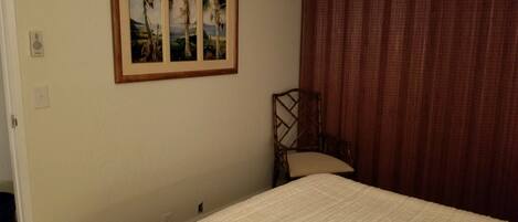 Un dormitorio con cama, una silla, un cuadro enmarco de palmeras y una ventana con cortinas.