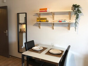Een eethoek met een witte tafel, zwarte stoelen en een spiegel aan de muur. Op de planken staan boeken en een geel modelbusje.