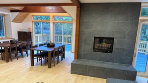 A spacious living area with a fireplace, wooden dining table, and large windows offering views of a deck and greenery. - Picollo Mondo (Yamanakako)
