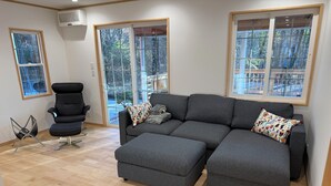 A modern living room with a grey sectional sofa, a black office chair, and a small side table. There are large windows with a view of trees and a deck. - Picollo Mondo (Yamanakako)