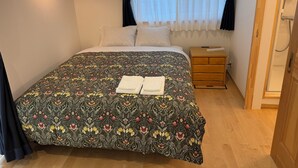 A neatly made bed with a floral bedspread, a bedside table with a remote control, and a window with sheer curtains. - Picollo Mondo (Yamanakako)