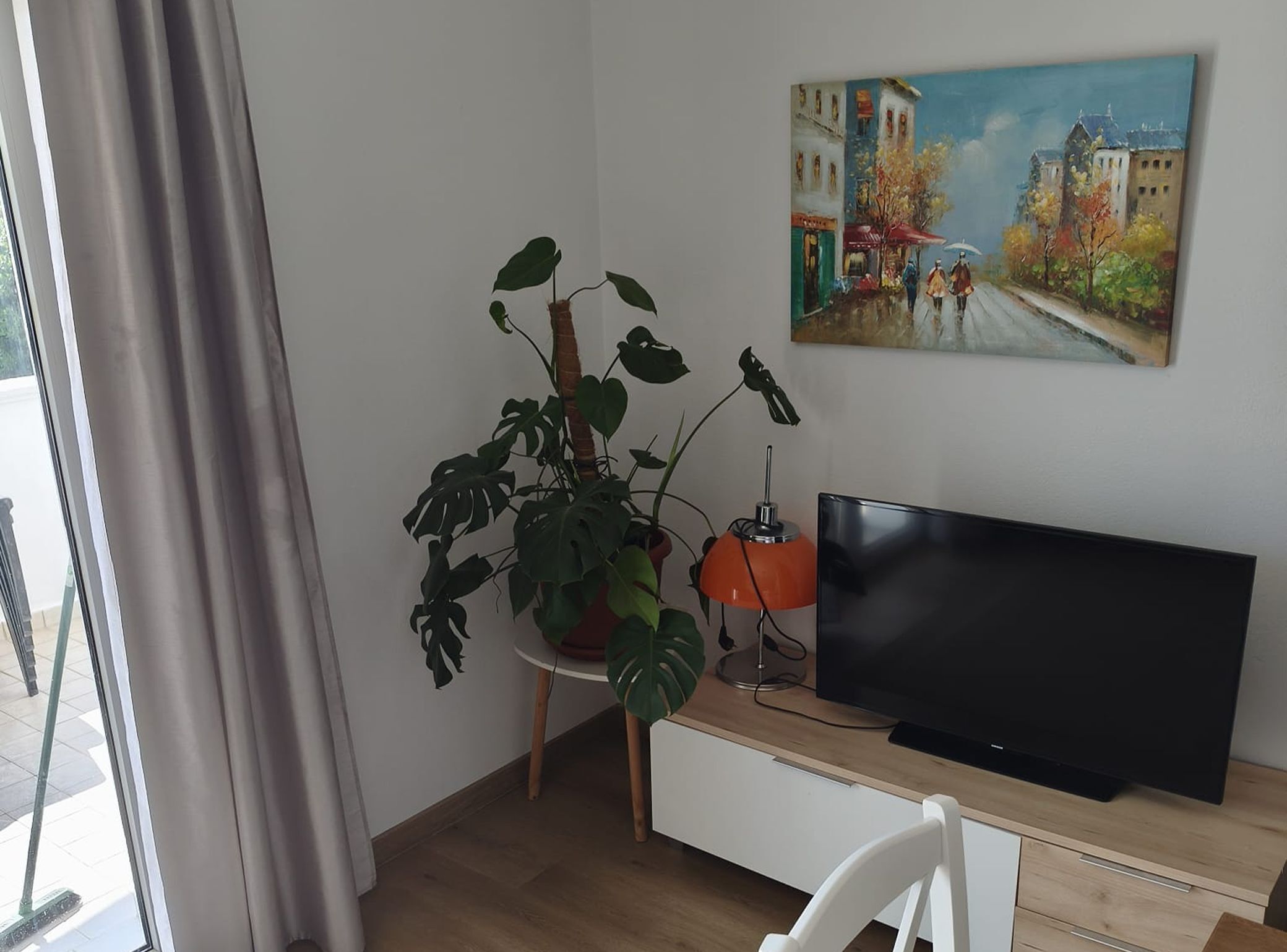 A room with a large window, a potted plant, a flat-screen TV, and a painting on the wall.