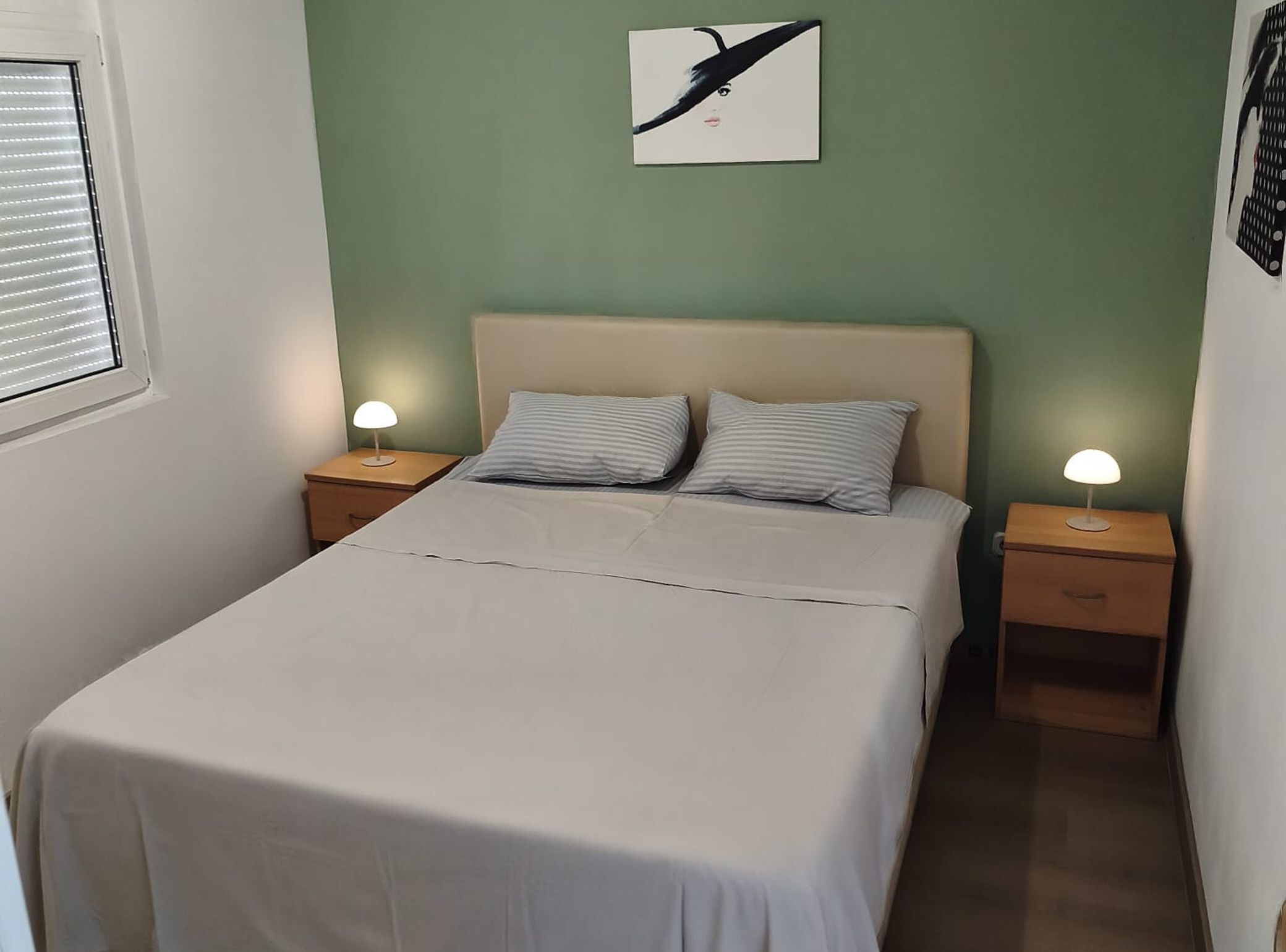 Un lit bien fait, recouvert d’une couette blanche et agrémenté de deux oreillers, est encadré de deux tables de chevet sur lesquelles reposent des lampes. La pièce est dotée de murs verts et d’une fenêtre avec des stores.