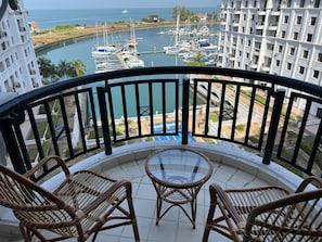 A balcony with wicker chairs and a glass table overlooking a marina with boats and a coastal view.