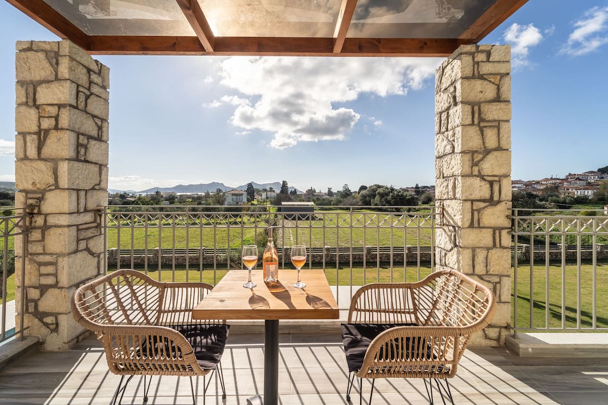 A covered outdoor seating area with a table and wicker chairs, overlooking a vineyard and distant hills.