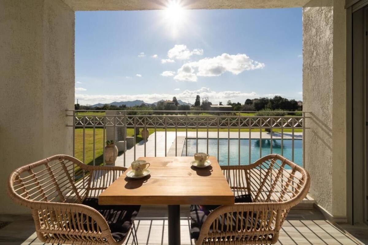 A balcony with wicker chairs, a wooden table, and a view of a swimming pool and landscape.