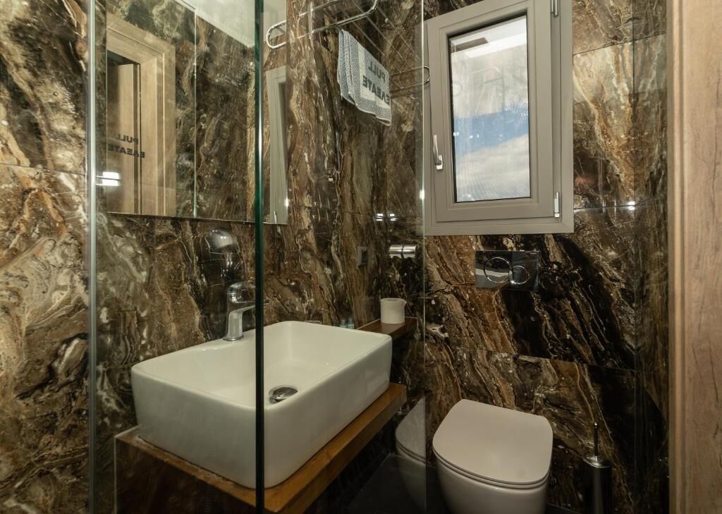 A bathroom with a marble wall, a white sink, a mirror, and a toilet.