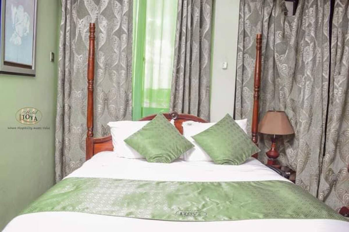Habitación de hotel con una cama, almohadas verdes, cabecera de madera, mesita de noche con una lámpara y cortinas estampadas.