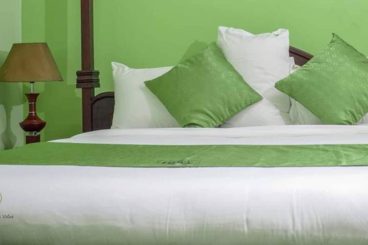 Una cama con ropa de cama blanca, almohadas verdes, un cabecero de madera, una lámpara y una pared verde.