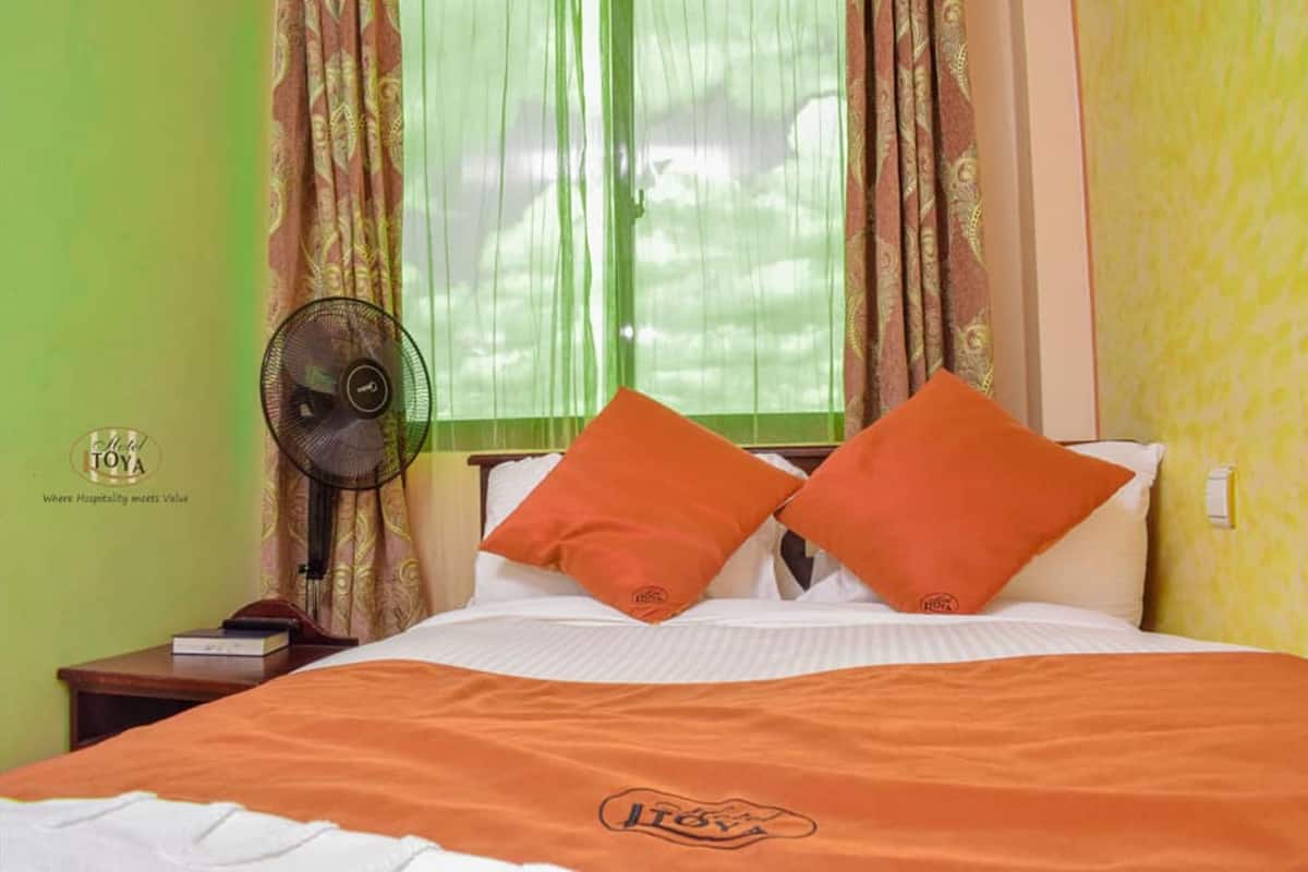 Una habitación de hotel con una cama, una colcha naranja, dos almohadas naranjas, un ventilador de noche y una ventana con cortinas traslúcidas.