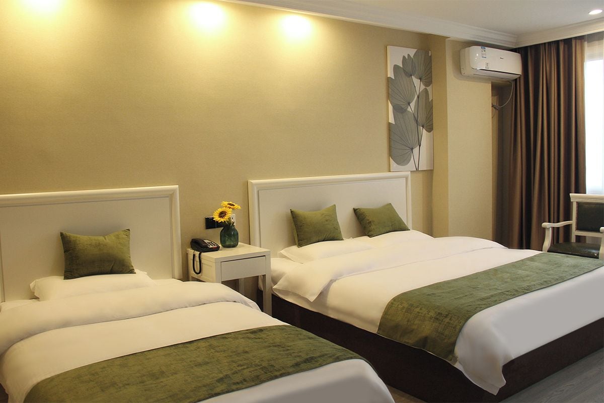 A hotel room with two beds, a bedside table with a lamp, and a wall-mounted air conditioner.