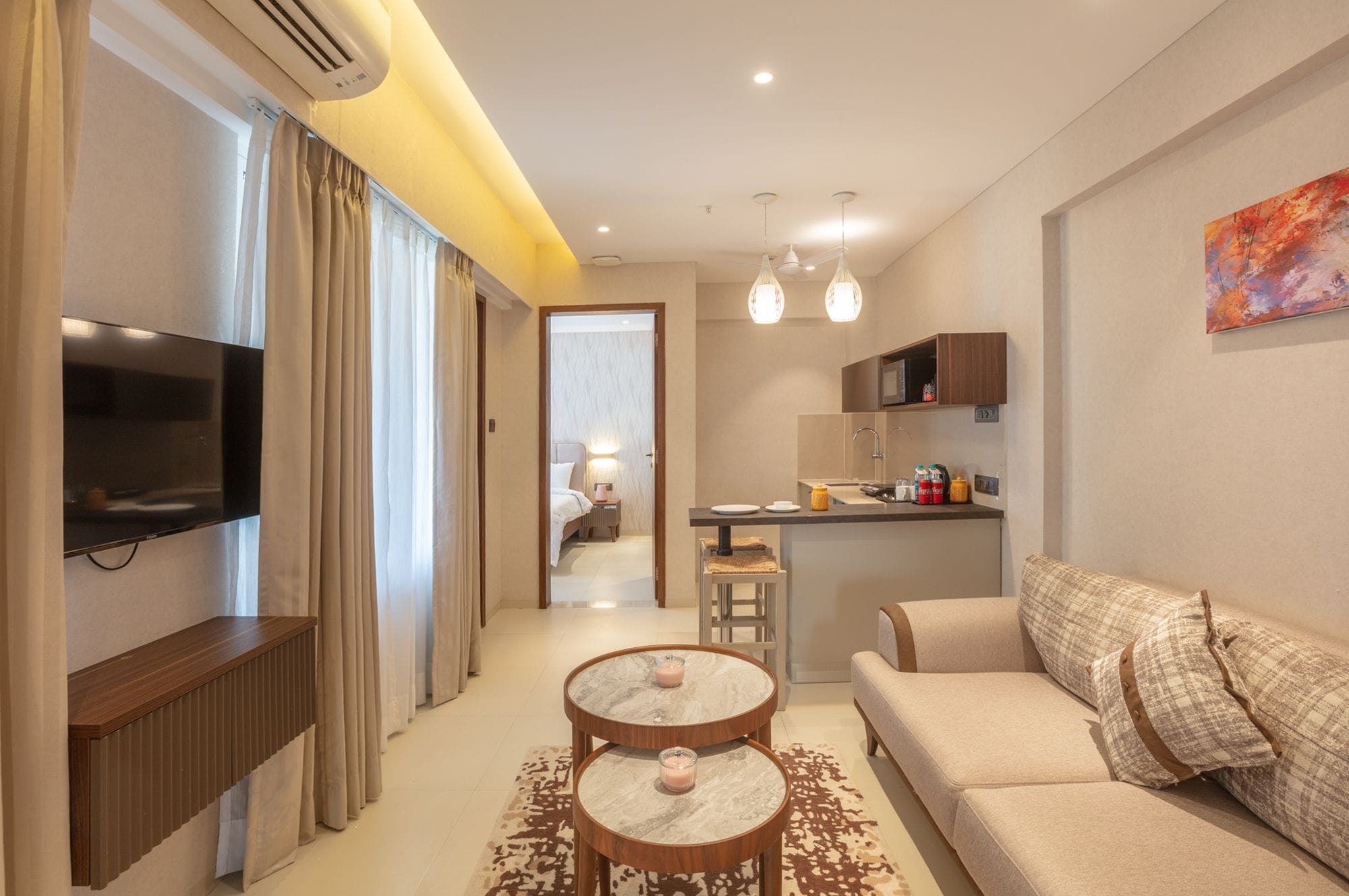 A modern hotel room with a TV, a sofa, a dining area, and a kitchen.