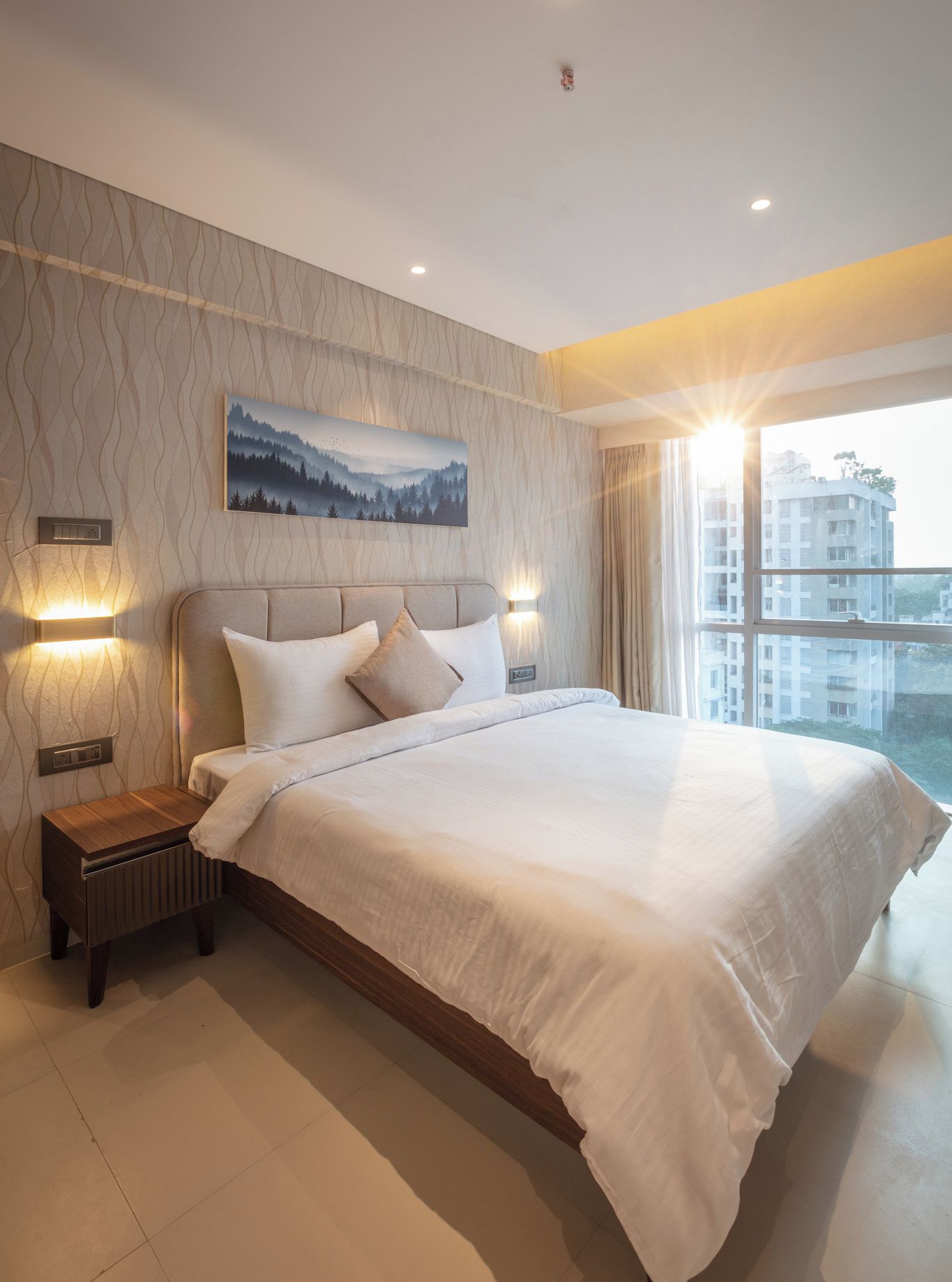 A modern bedroom with a large bed, two bedside lamps, a nightstand, a wall-mounted painting, and a large window with a view of buildings.