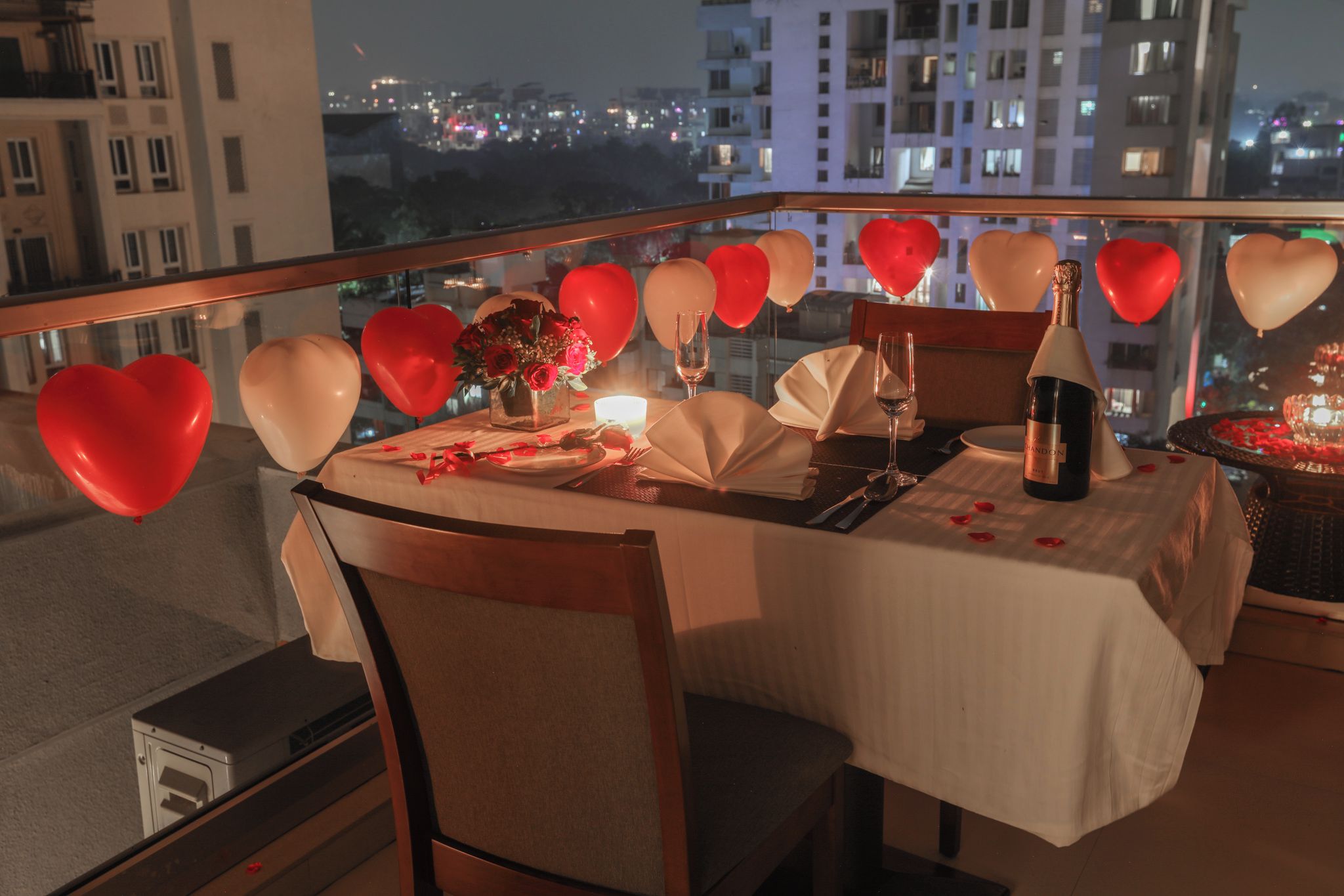 A table set for two with a bottle of champagne, a vase of flowers, and heart-shaped balloons.