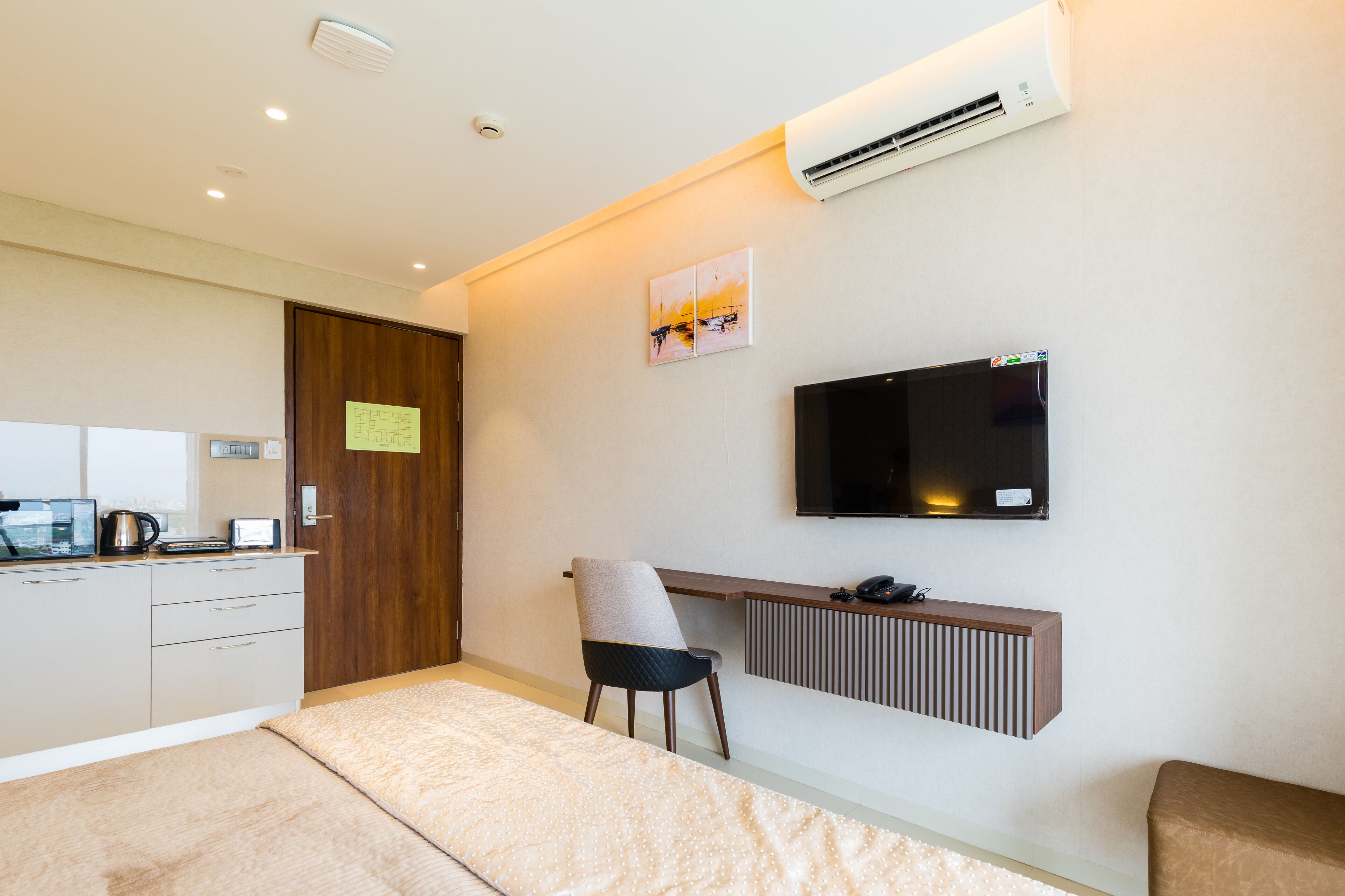 A modern hotel room with a bed, a desk with a chair, a television, and an air conditioning unit.