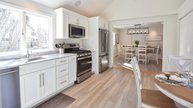 Una cocina moderna con armarios blancos, electrodomésticos de acero inoxidable y un comedor con mesa y sillas de madera.