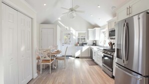 Una cocina con armarios blancos, electrodomésticos de acero inoxidable, un ventilador de techo y un comedor con sillas de madera y una mesa.