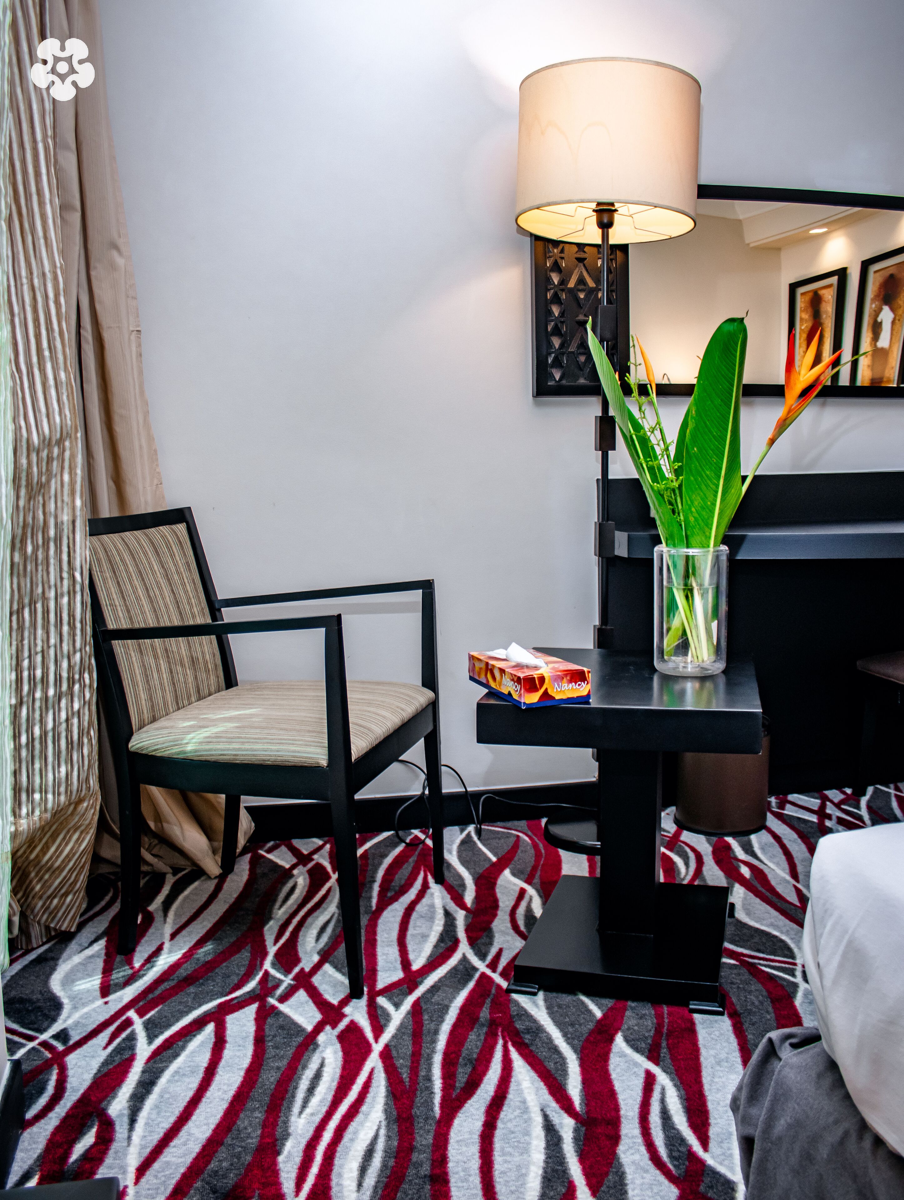 A hotel room with a bed, a chair, a desk with a lamp, a vase with plants, and a patterned rug.