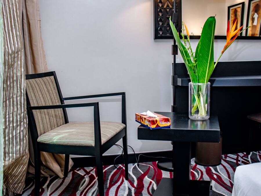 A hotel room with a bed, a chair, a desk with a lamp, a vase with plants, and a patterned rug.