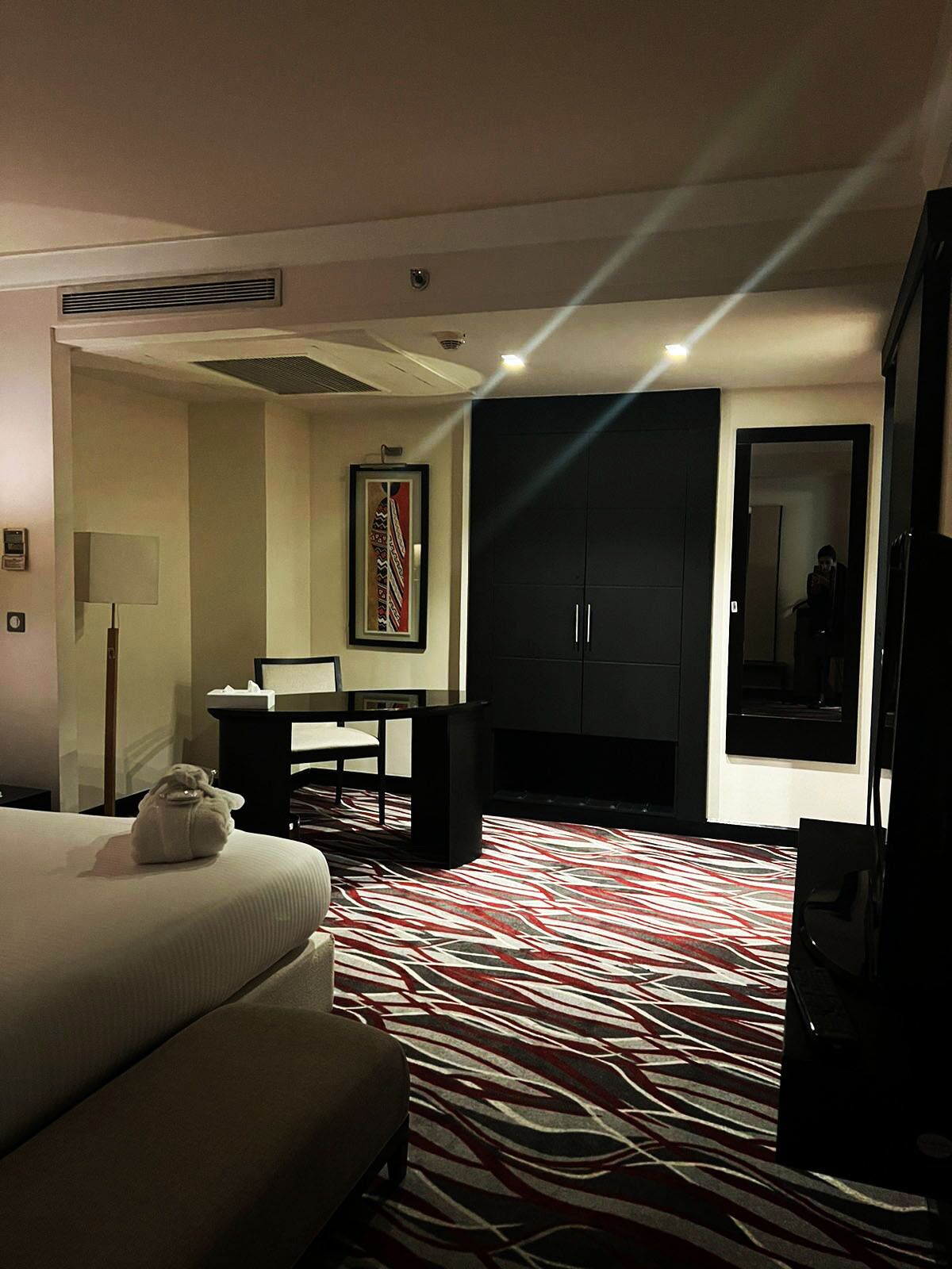 A hotel room with a bed, a desk, a chair, a wardrobe, and a large mirror.