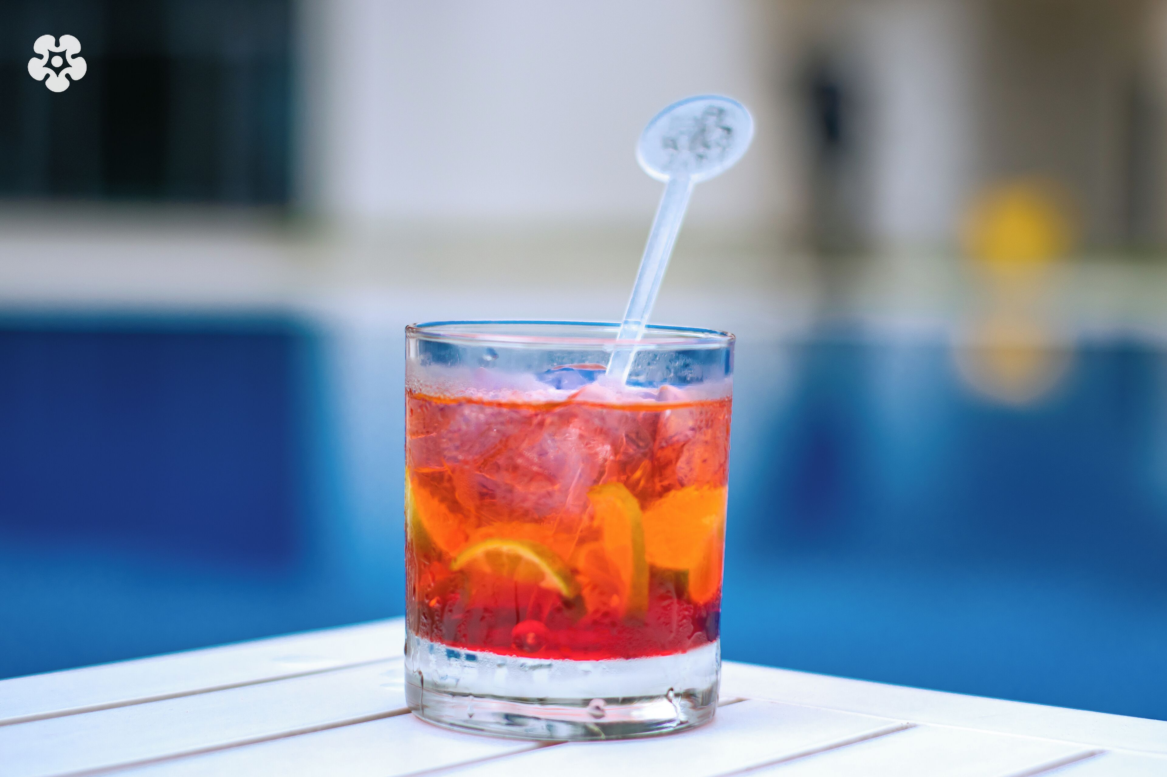 A glass with a clear liquid, ice, and orange slices, placed on a white surface near a pool.