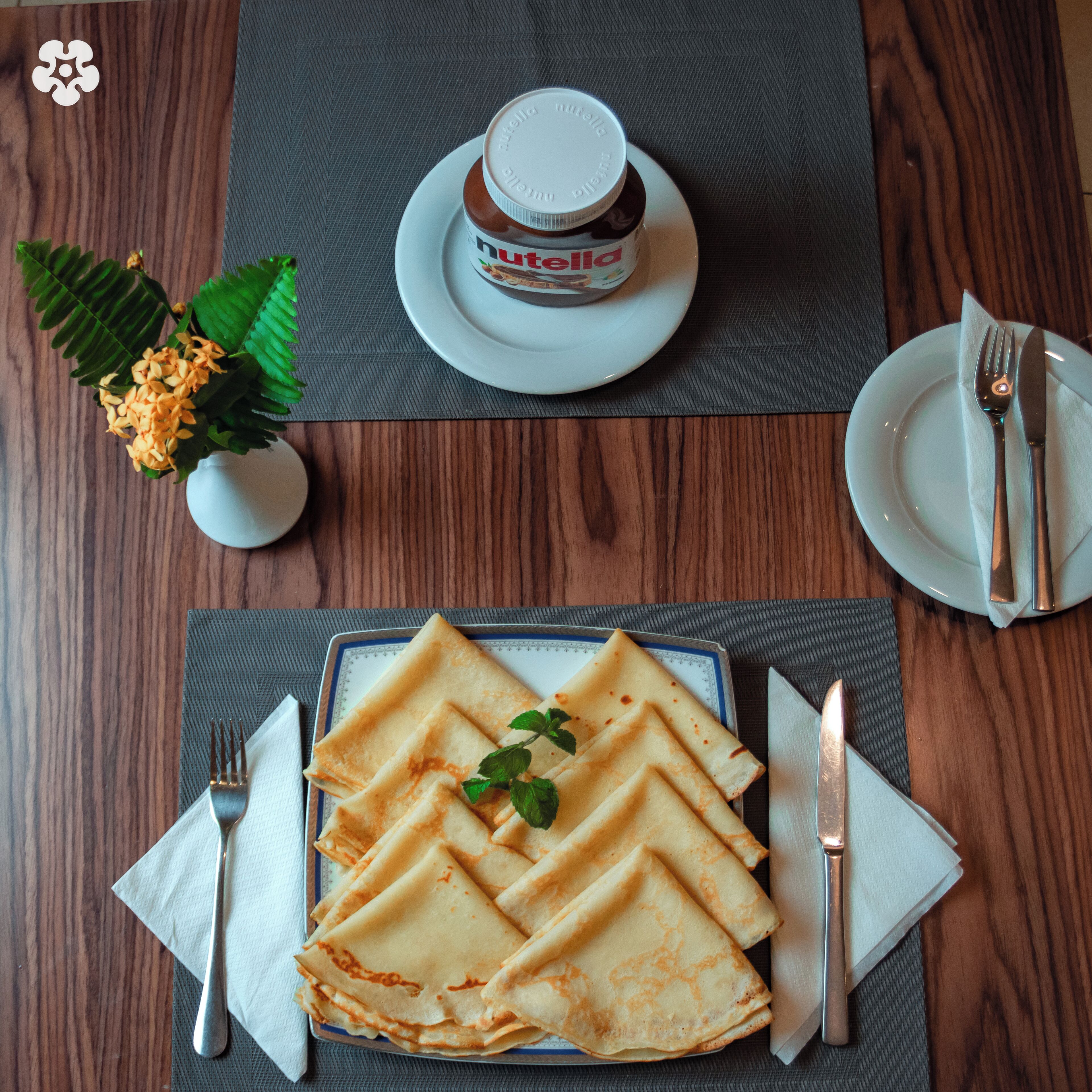 A dining table with a plate of crepes, a Nutella jar, and a small vase with flowers.