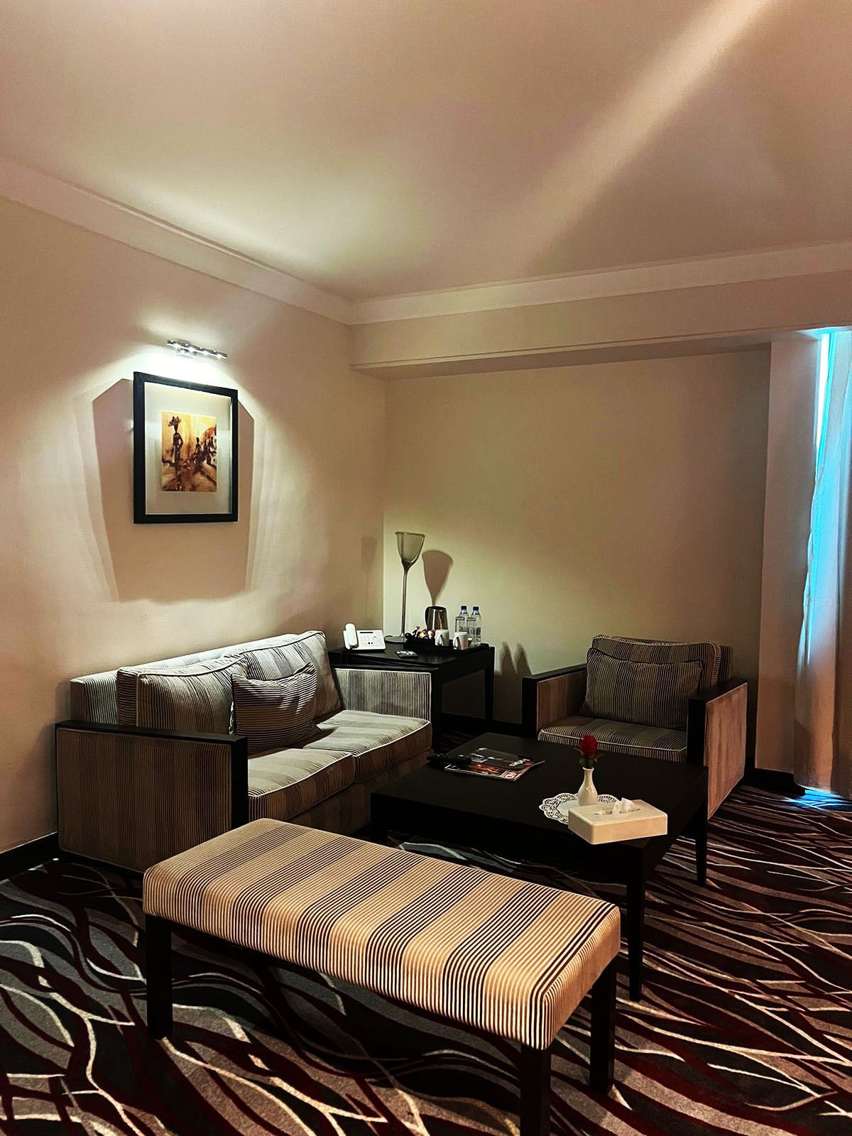 A hotel room with a sofa, armchair, and bench, a small table with a lamp and a tray, and a framed picture on the wall.