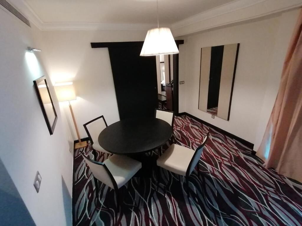 Ett hotellrum med ett runt bord, två stolar, en hängande lampa, en mönstrad matta och en stor spegel på väggen.