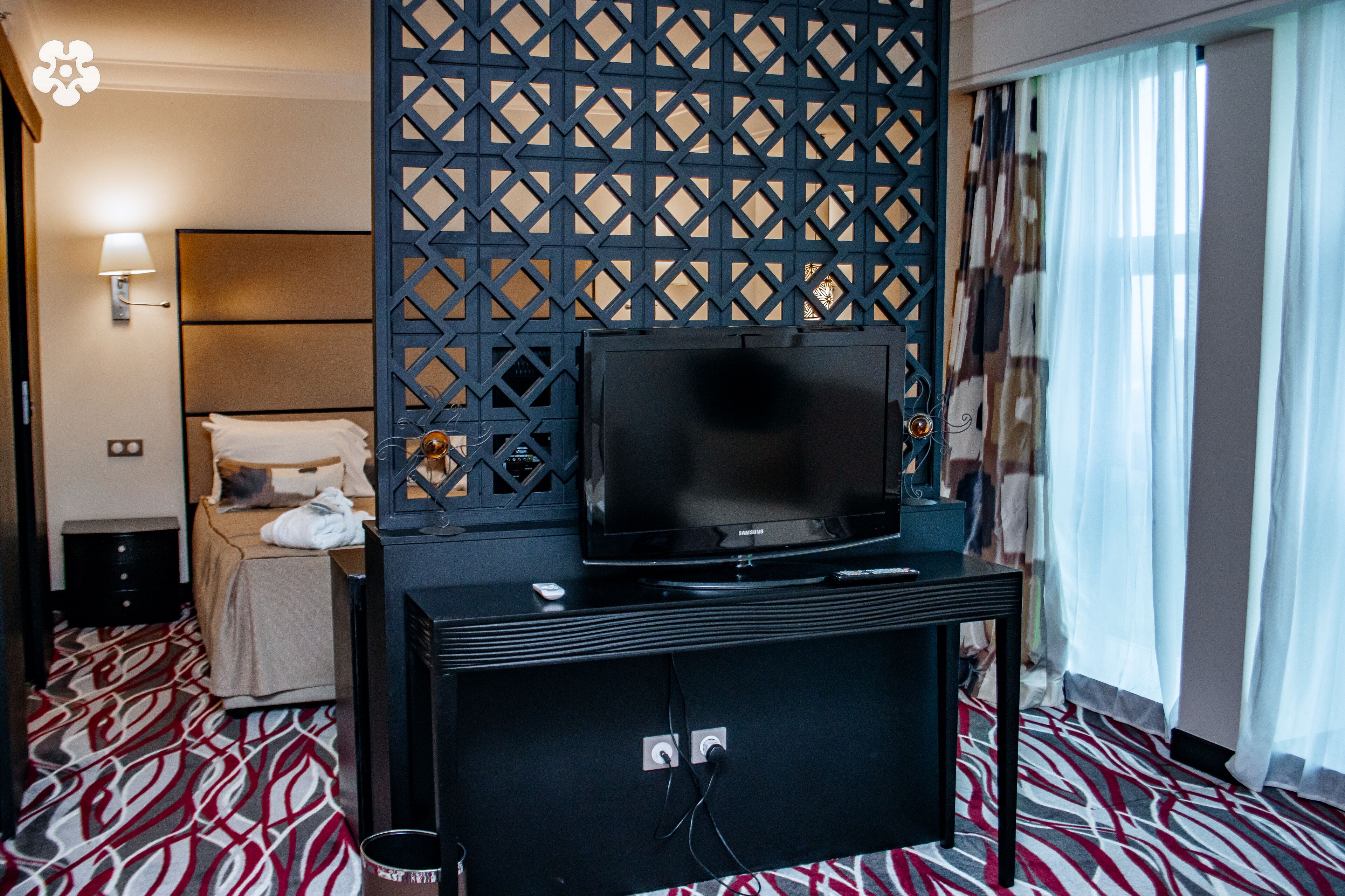 A hotel room with a bed, a TV on a stand, a wall-mounted TV, a patterned carpet, a nightstand, a lamp, and a window with curtains.