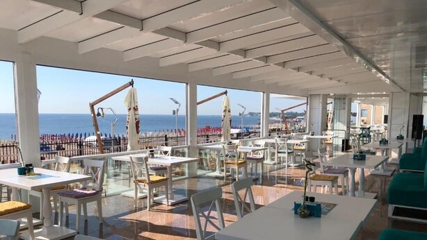 An outdoor dining area with tables and chairs, large windows overlooking the sea, and umbrellas. - Hotel Pasha (Eforie)